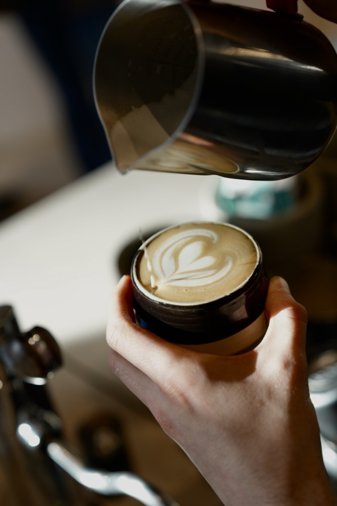 Eine Hand gießt Milch in einen Cappuccino, im Schaum entsteht eine Rosetta