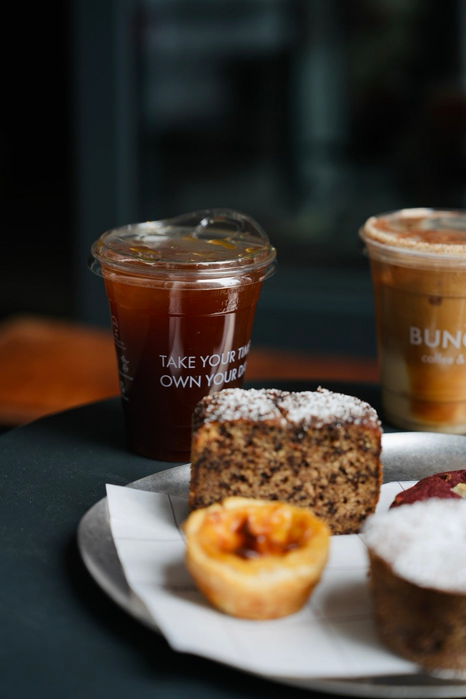 Cold Brew und Iced Latte im Becher, daneben ein Teller mit Kuchen und Gebäck