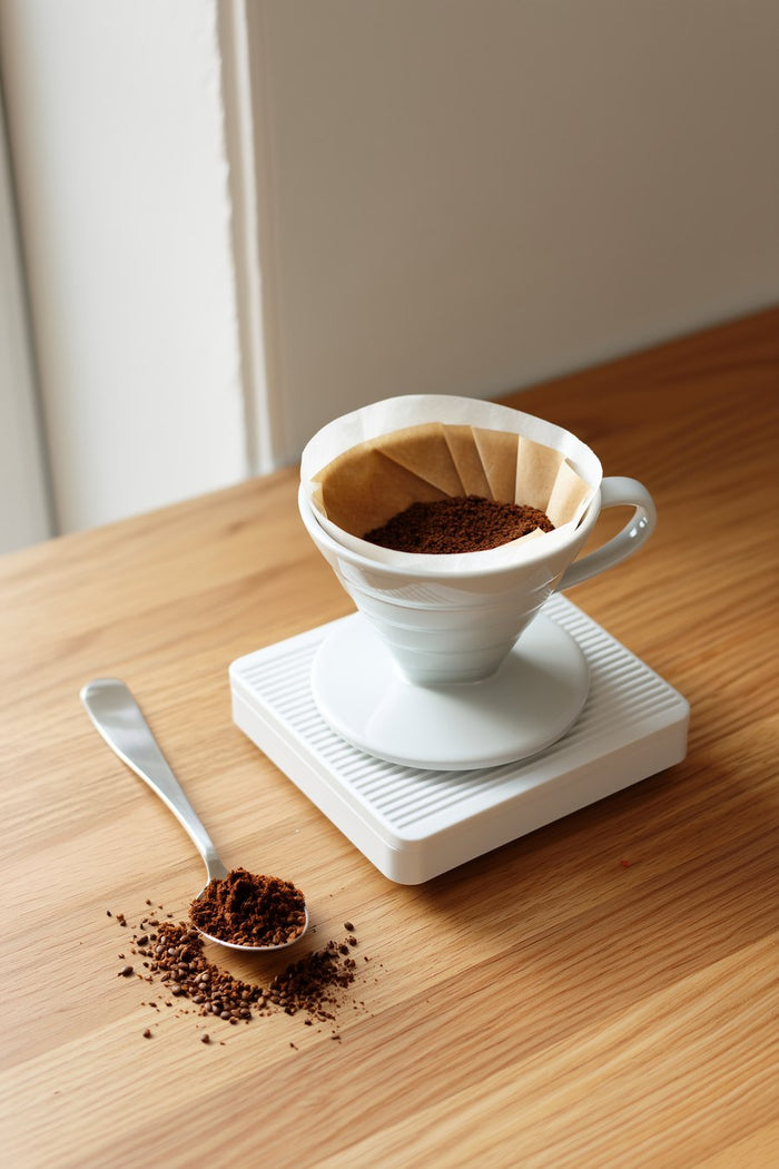 Handfilter mit gemahlenem Kaffee steht auf einer Küchenwaage, daneben ein gehäufter Esslöffel Kaffeemehl